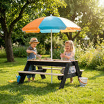 Two children playing at a sand table with a colorful umbrella in a garden.