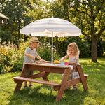 Two children playing at a wooden picnic table with a striped umbrella in a garden.